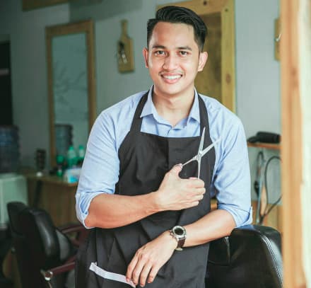 Barbeiro homem latino em frente a uma barbearia enconstado na porta com uma tesoura na mão e sorrindo